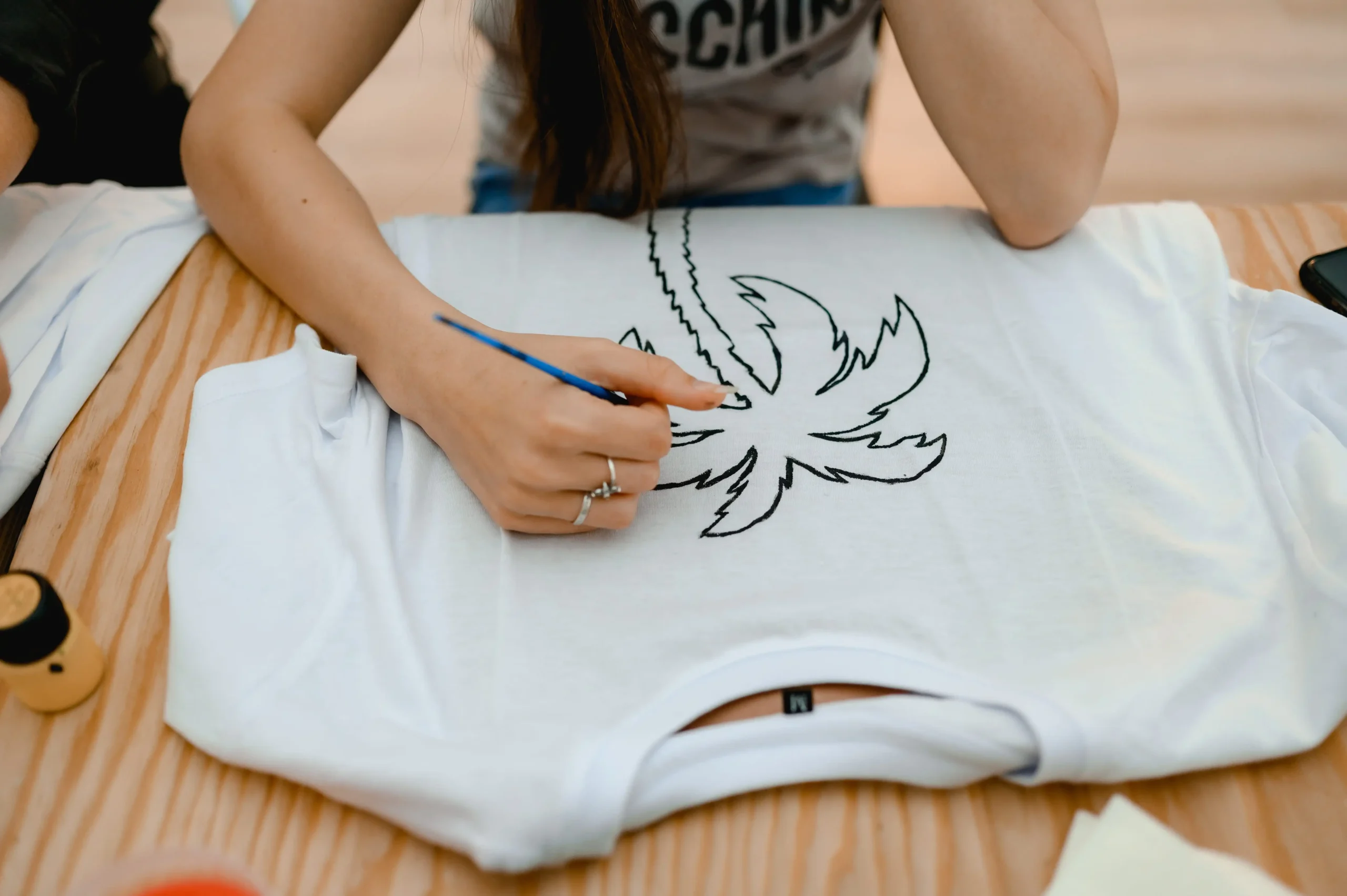 Can I Embroider Directly on a Shirt? Hands drawing a floral design on a white T-shirt laid flat on a wooden table.