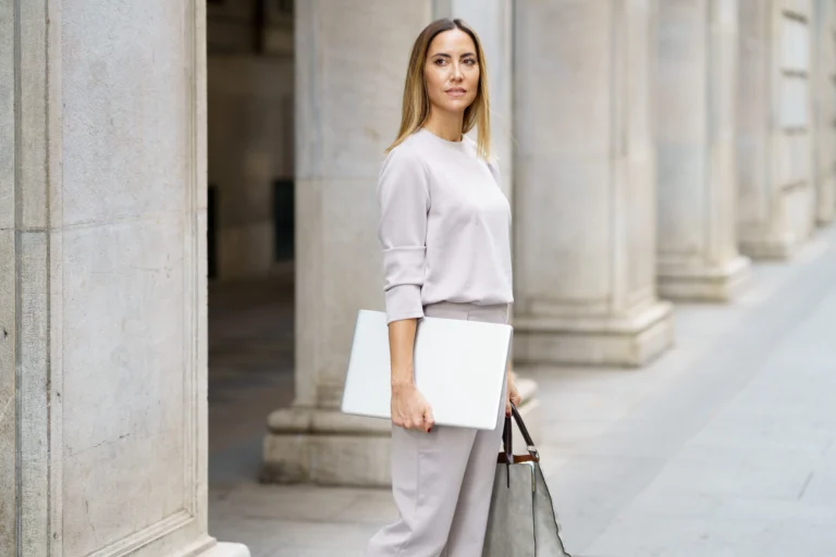 A professionally dressed woman stands near stone columns, holding a laptop and a handbag, looking off to the side.