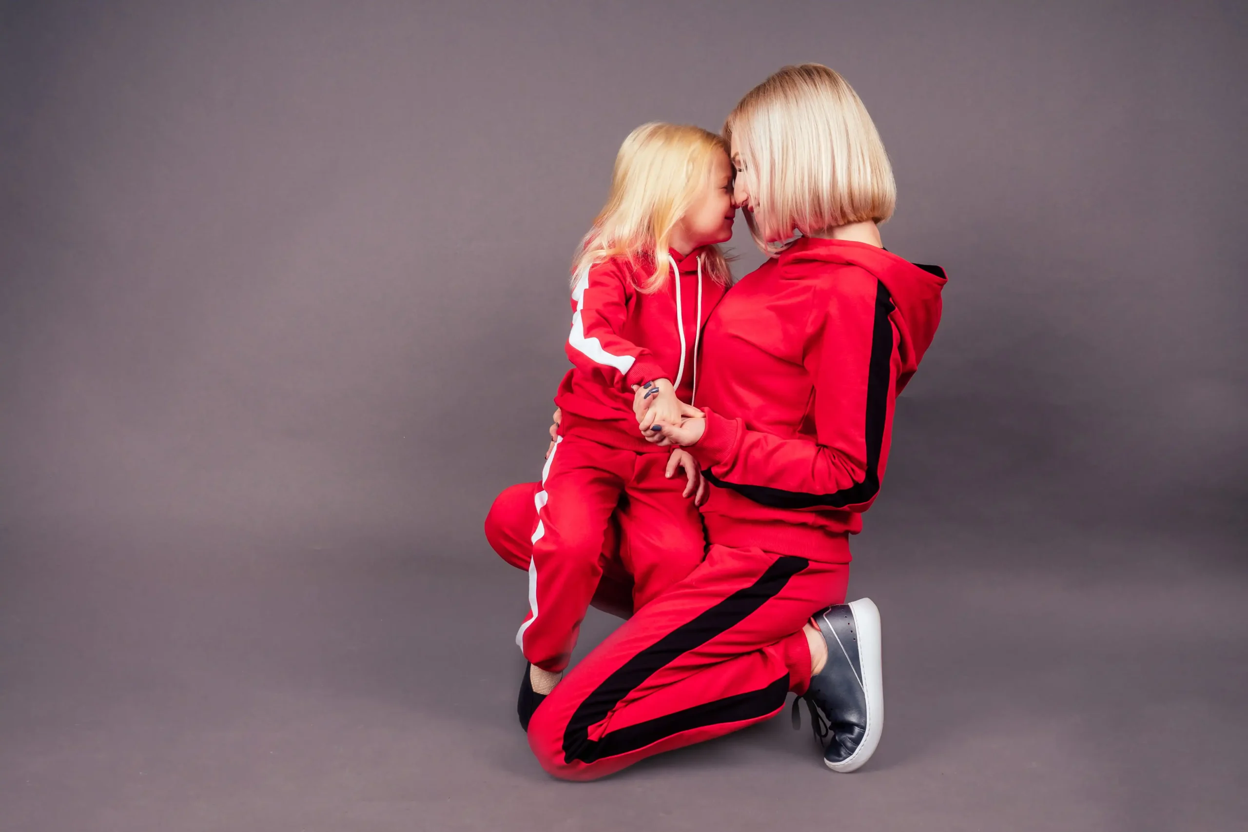 Woman and young girl in matching red tracksuits kneel facing each other and hold hands against a gray background.