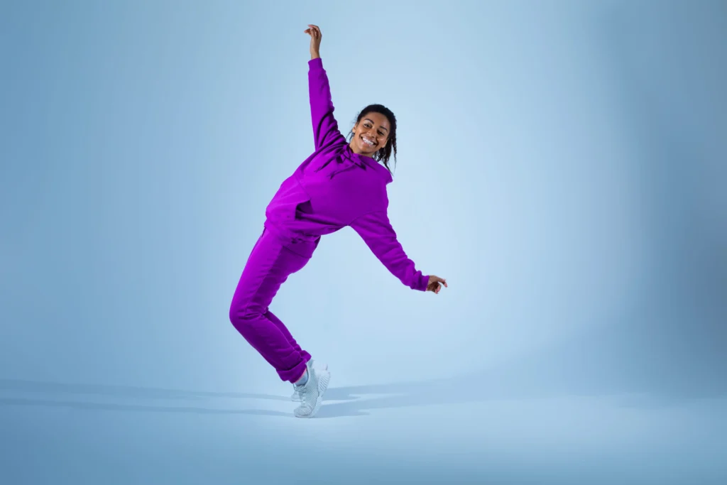 Smiling woman in a bright purple tracksuit posing playfully with one arm raised against a light blue background.
