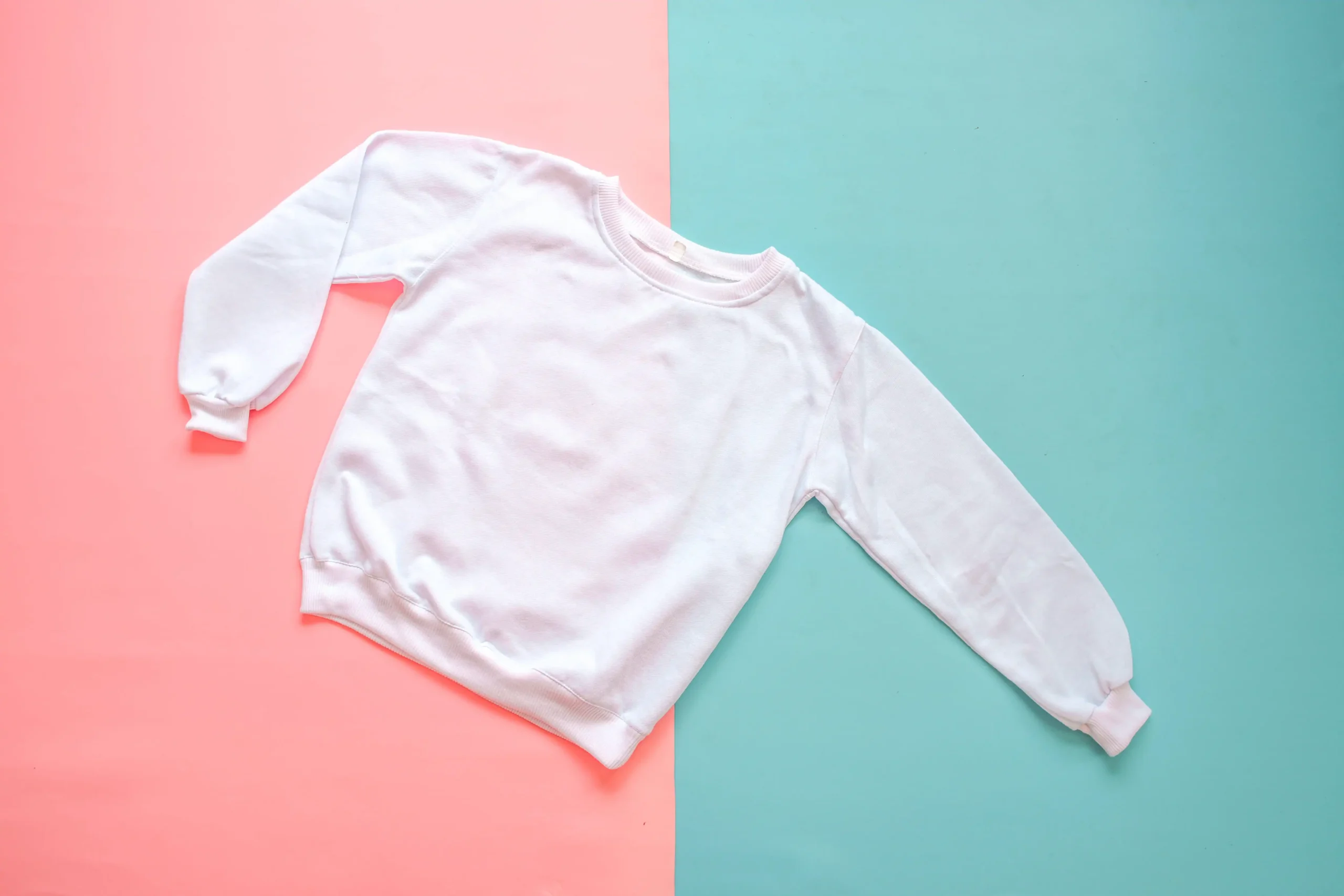 Plain white sweatshirt laid flat on a split pastel background of pink and teal.