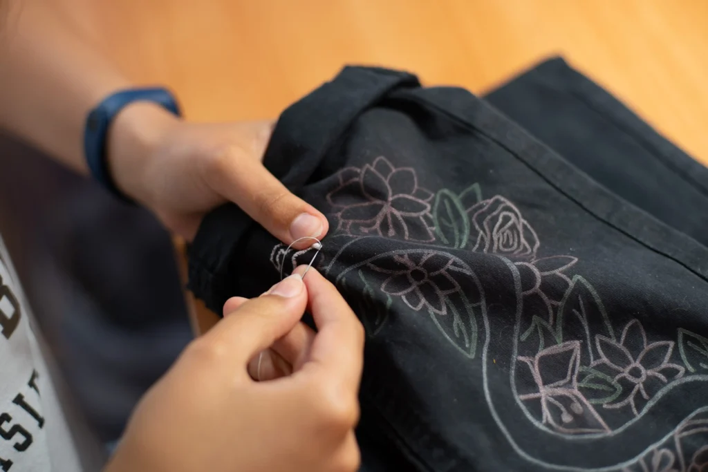 Hands carefully embroidering a floral pattern onto black fabric using a needle and thread.